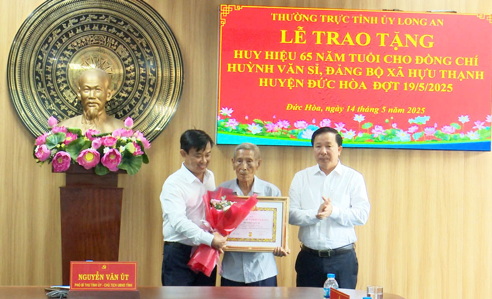 Phó Bí thư Tỉnh ủy, Chủ tịch UBND tỉnh - Nguyễn Văn Út (bên phải) và Bí thư Huyện ủy Đức Hòa - Nguyễn Minh Hùng trao Huy hiệu 65 năm tuổi Đảng và tặng hoa chúc mừng đảng viên Huỳnh Văn Sỉ